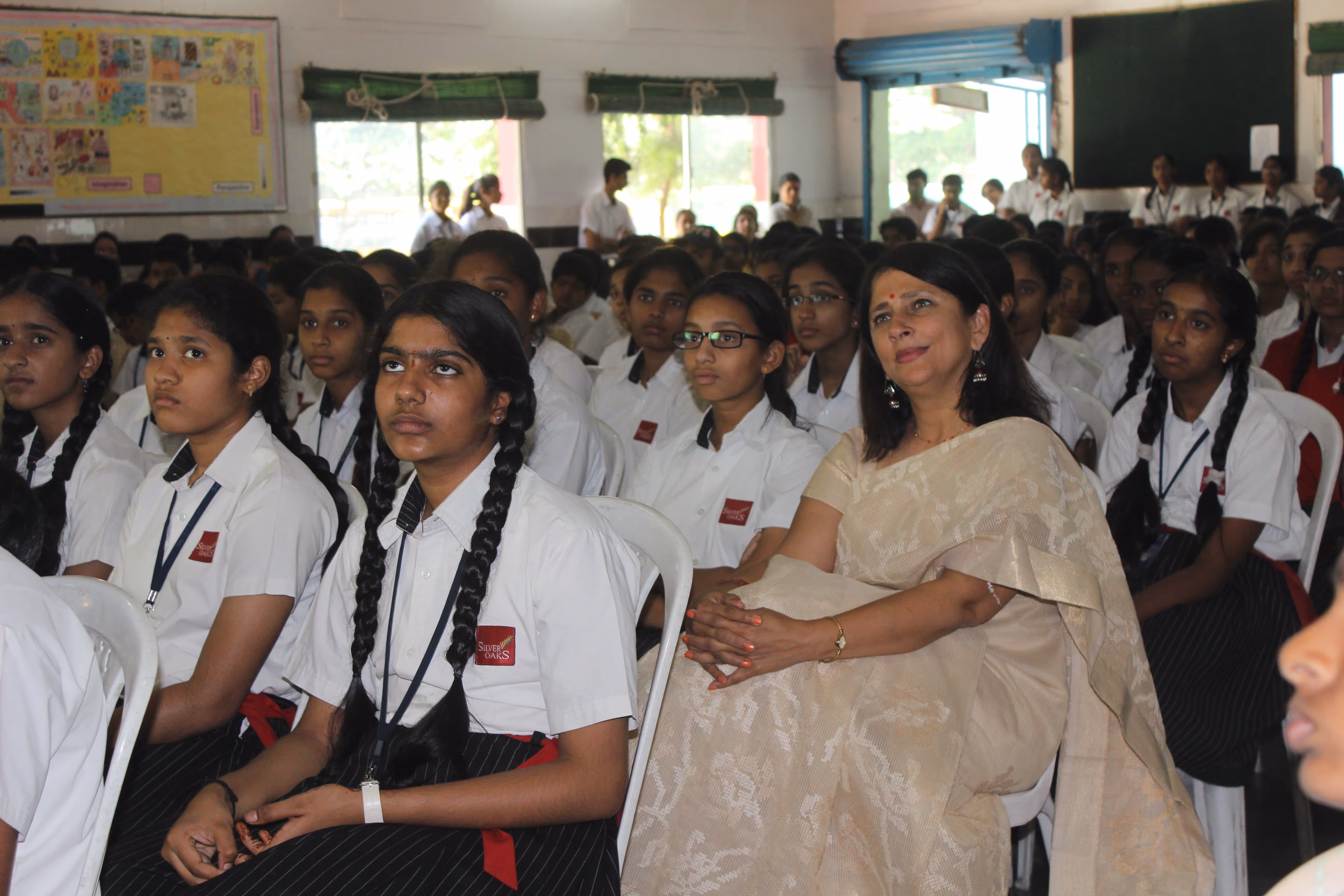 Group of female students in white uniforms sitting attentively with a woman in a beige saree in a classroom.