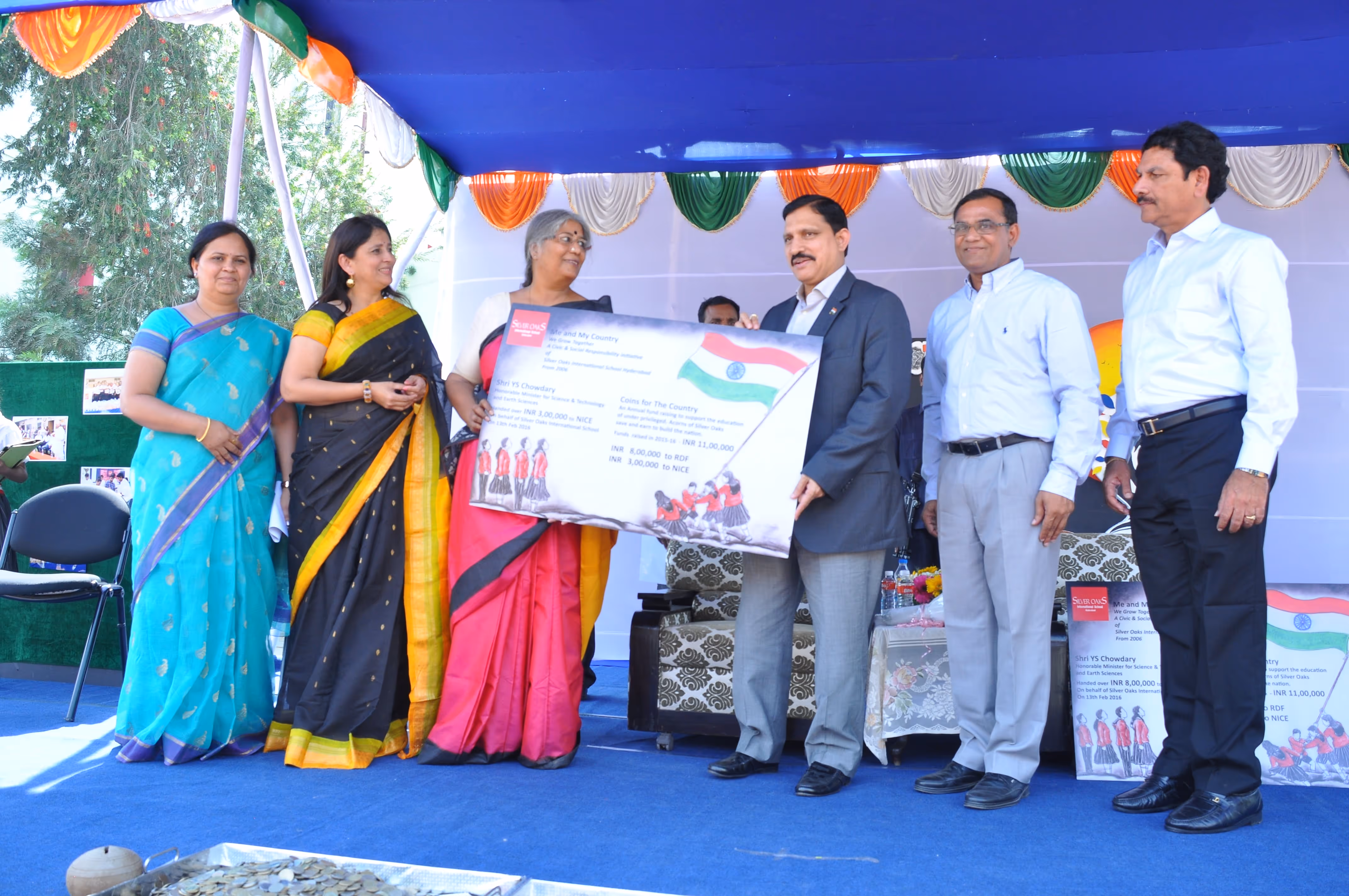 Group of six people standing on a stage under a canopy decorated with green, white, and orange drapes, with two women holding a large ceremonial check featuring the Indian flag.