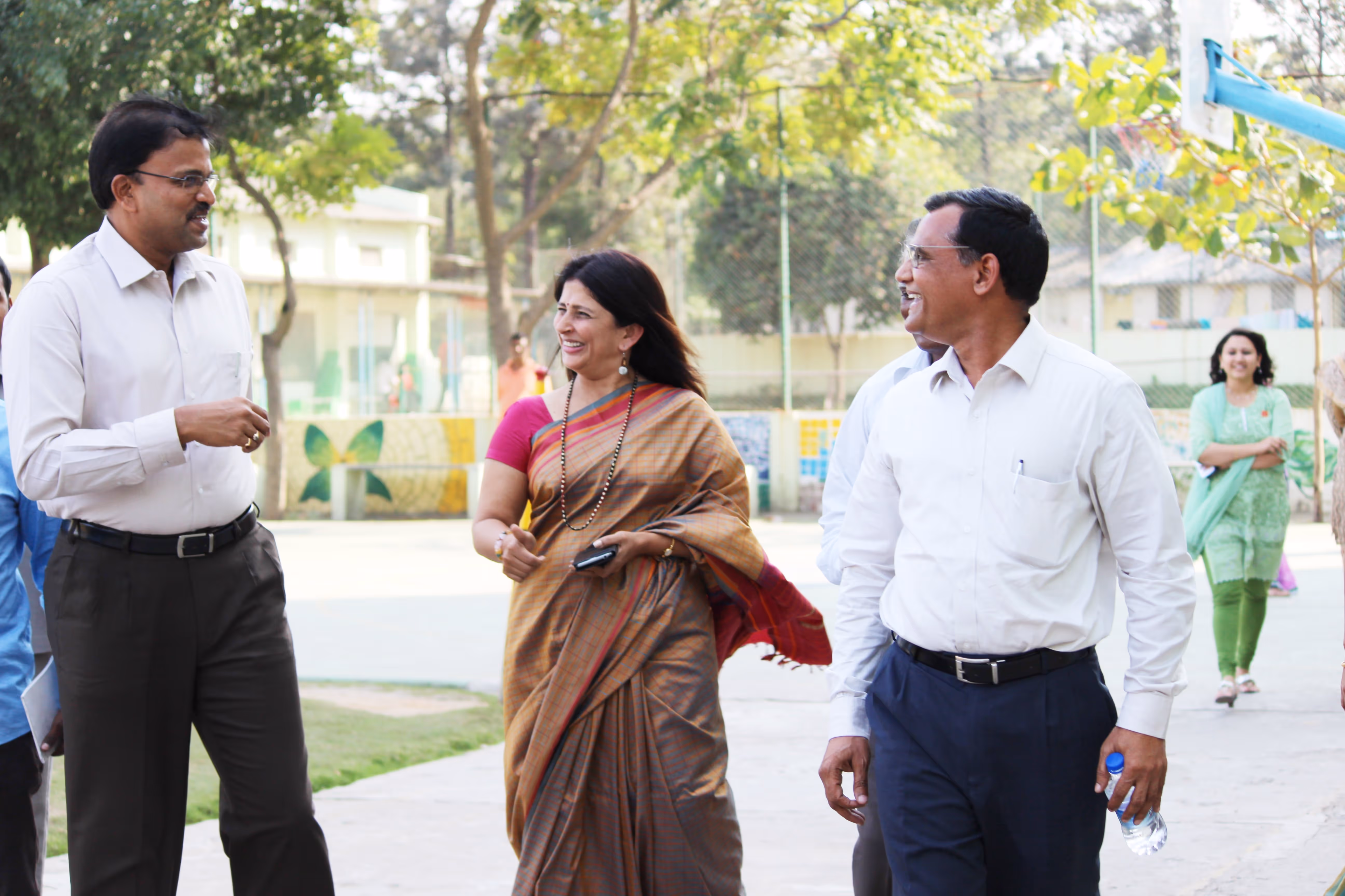 Three adults smiling and walking outdoors on a paved path with trees and a fence in the background.