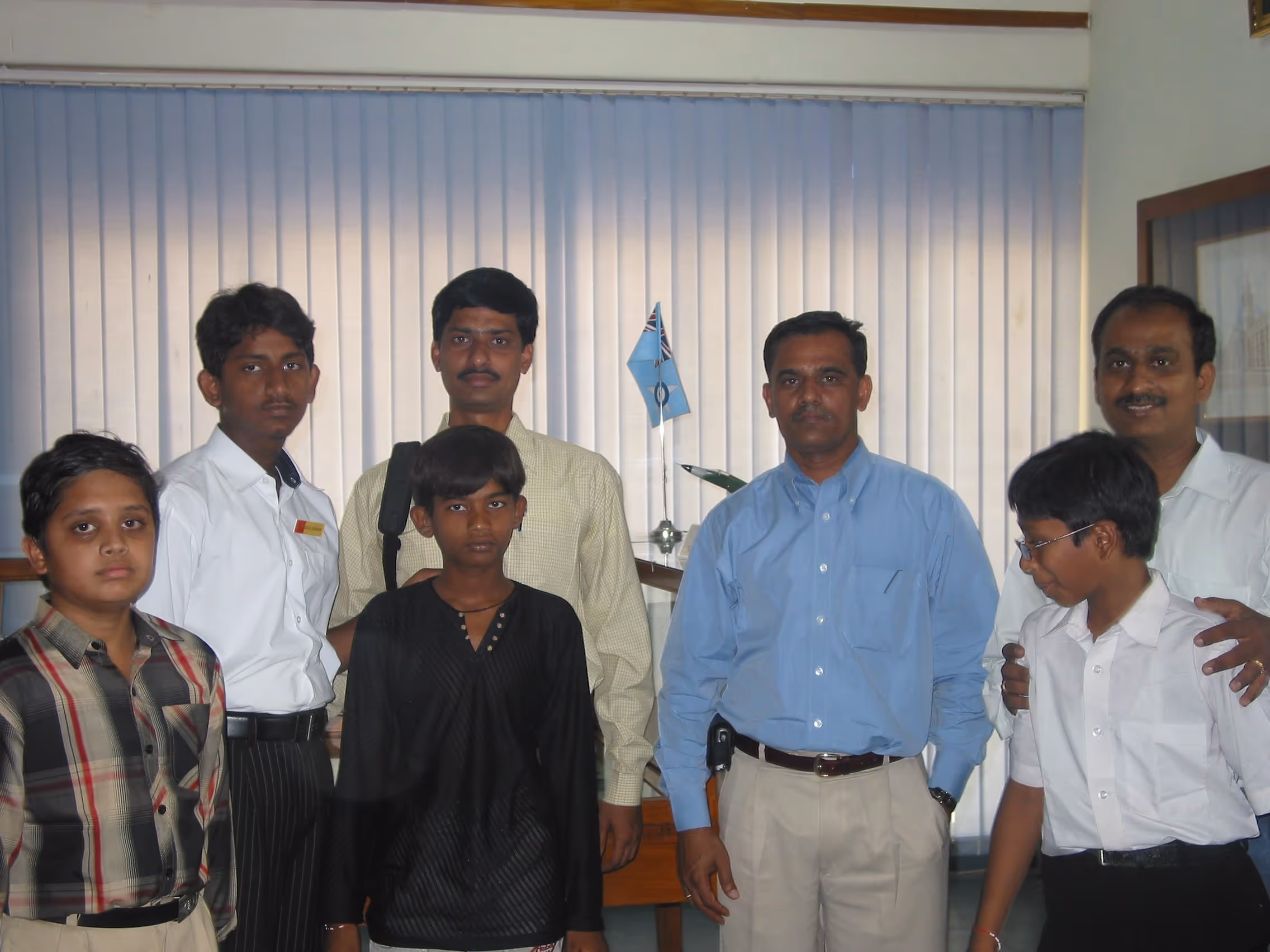 Group photo of four men and three boys standing indoors in front of vertical blinds and a desk with a small flag on it.