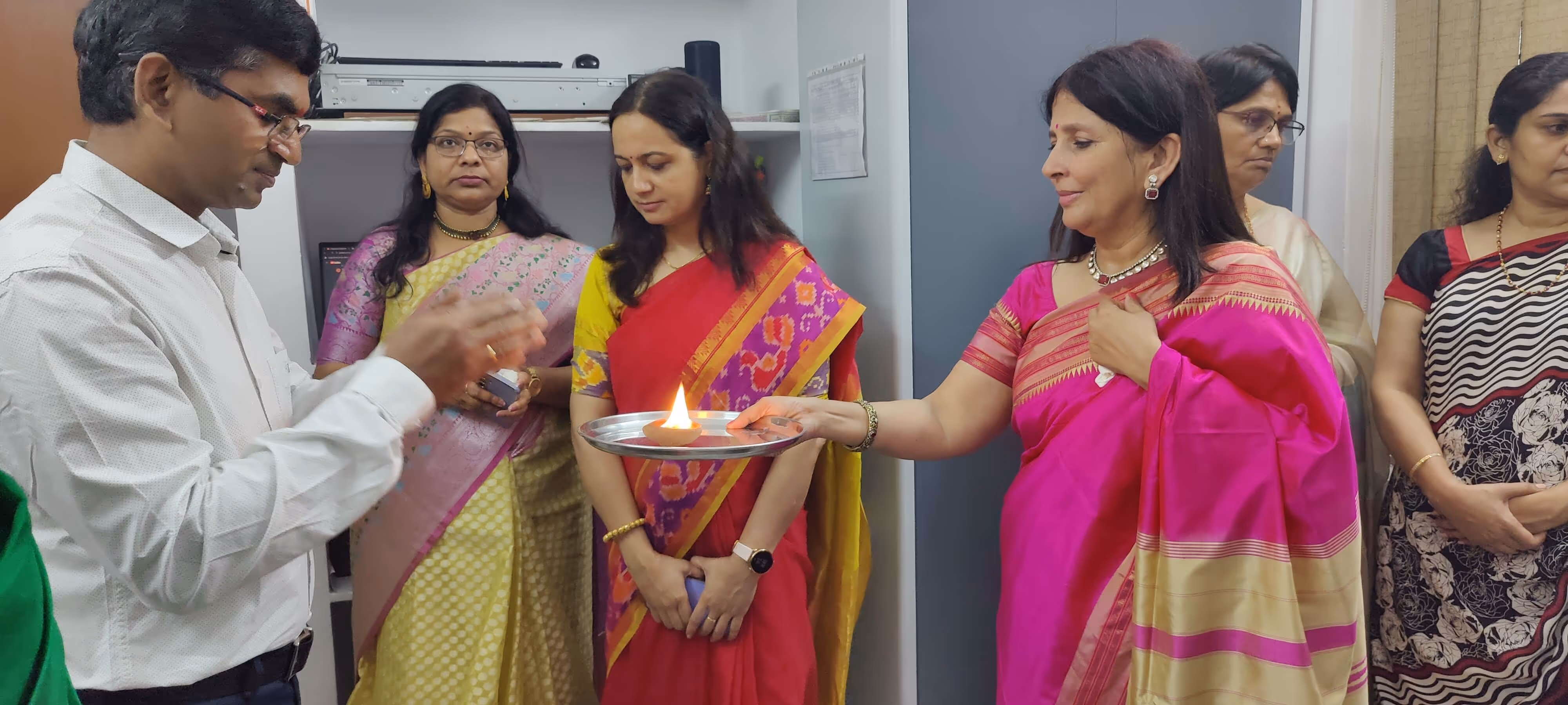Group of people in traditional Indian attire participating in a ritual with a lit oil lamp on a metal plate.