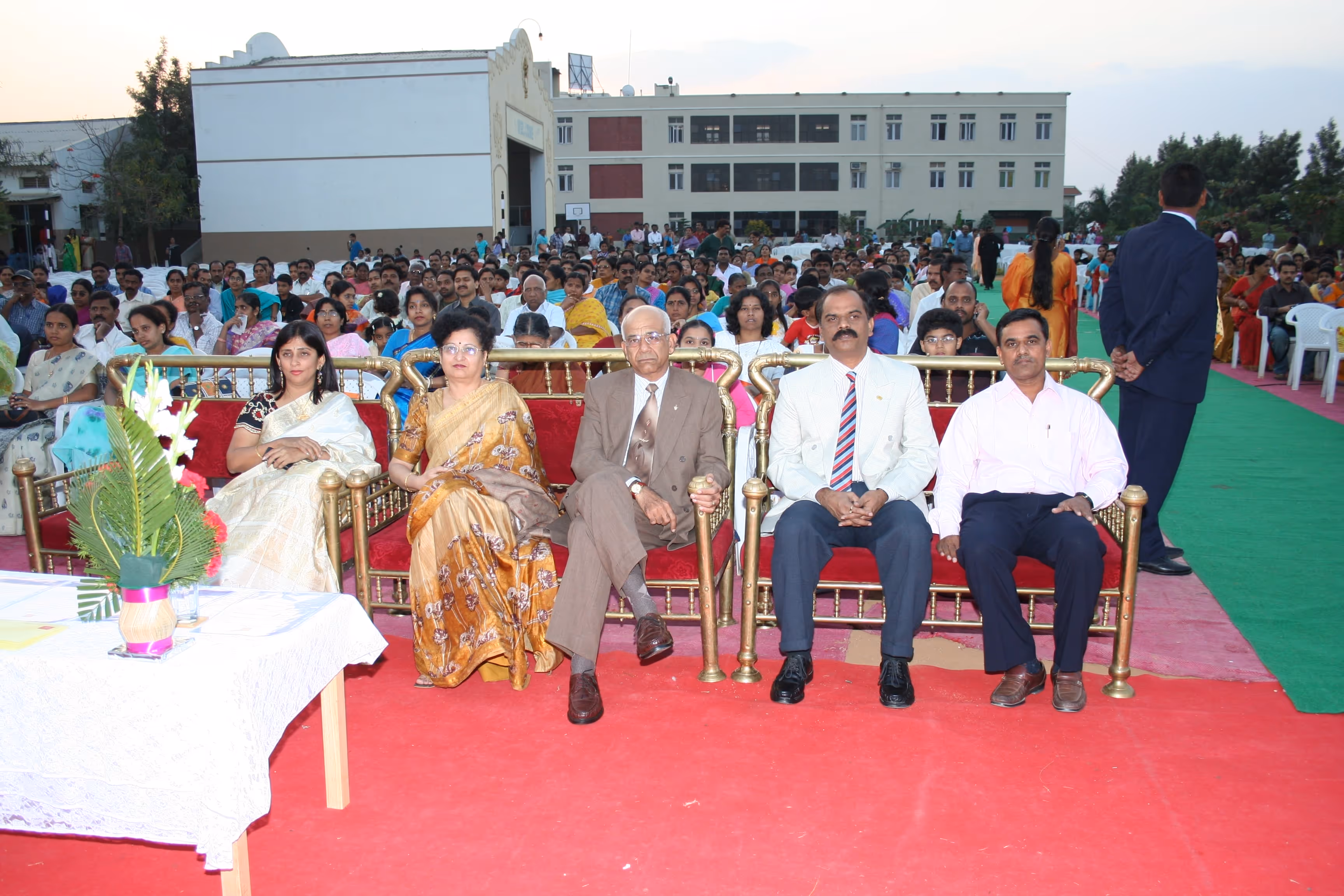 Five formally dressed individuals seated on a row of gold and red chairs in front of a large audience at an outdoor event.