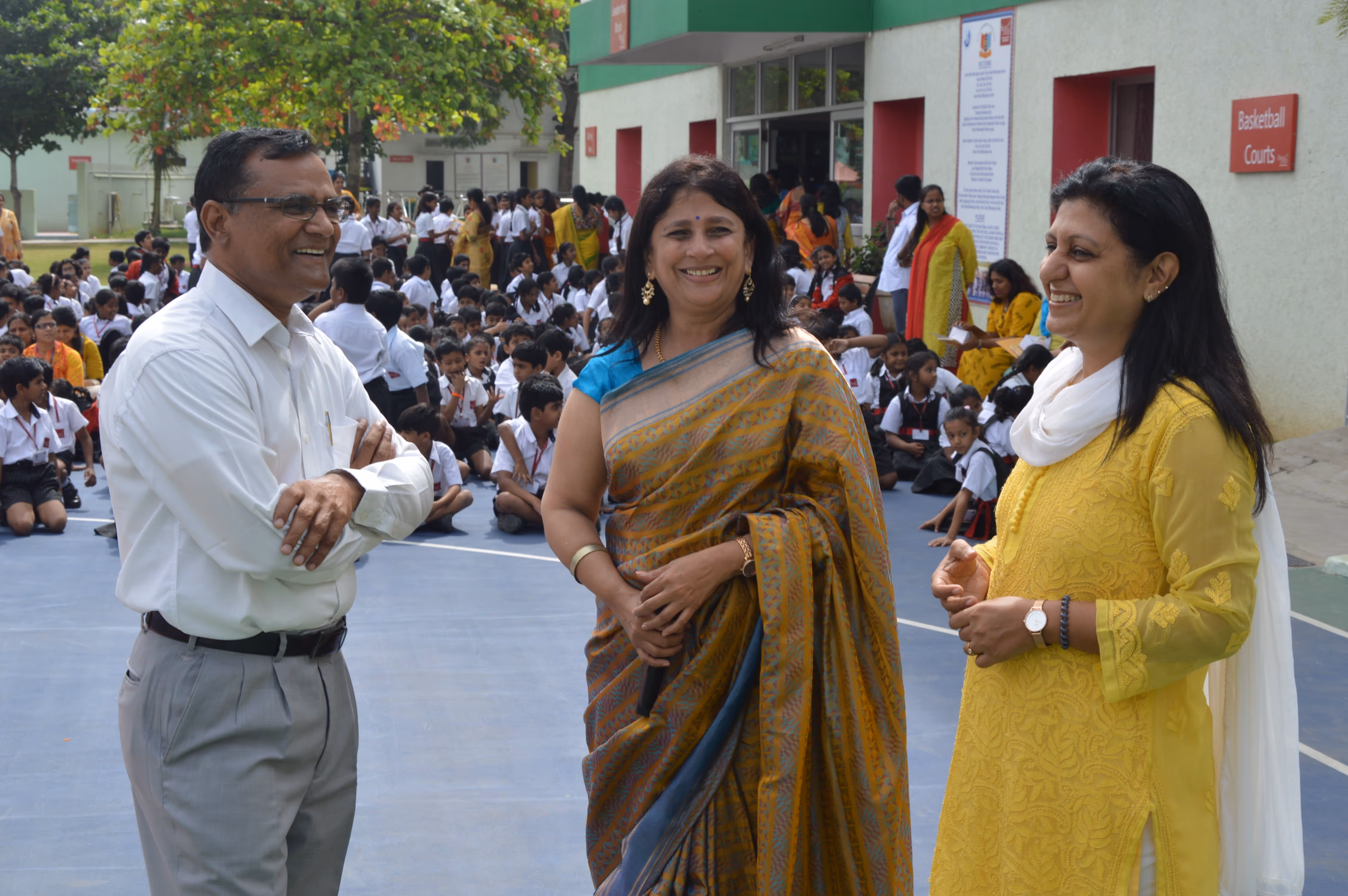 Three adults smiling and conversing outdoors on a basketball court with a large group of seated school children in uniforms in the background.