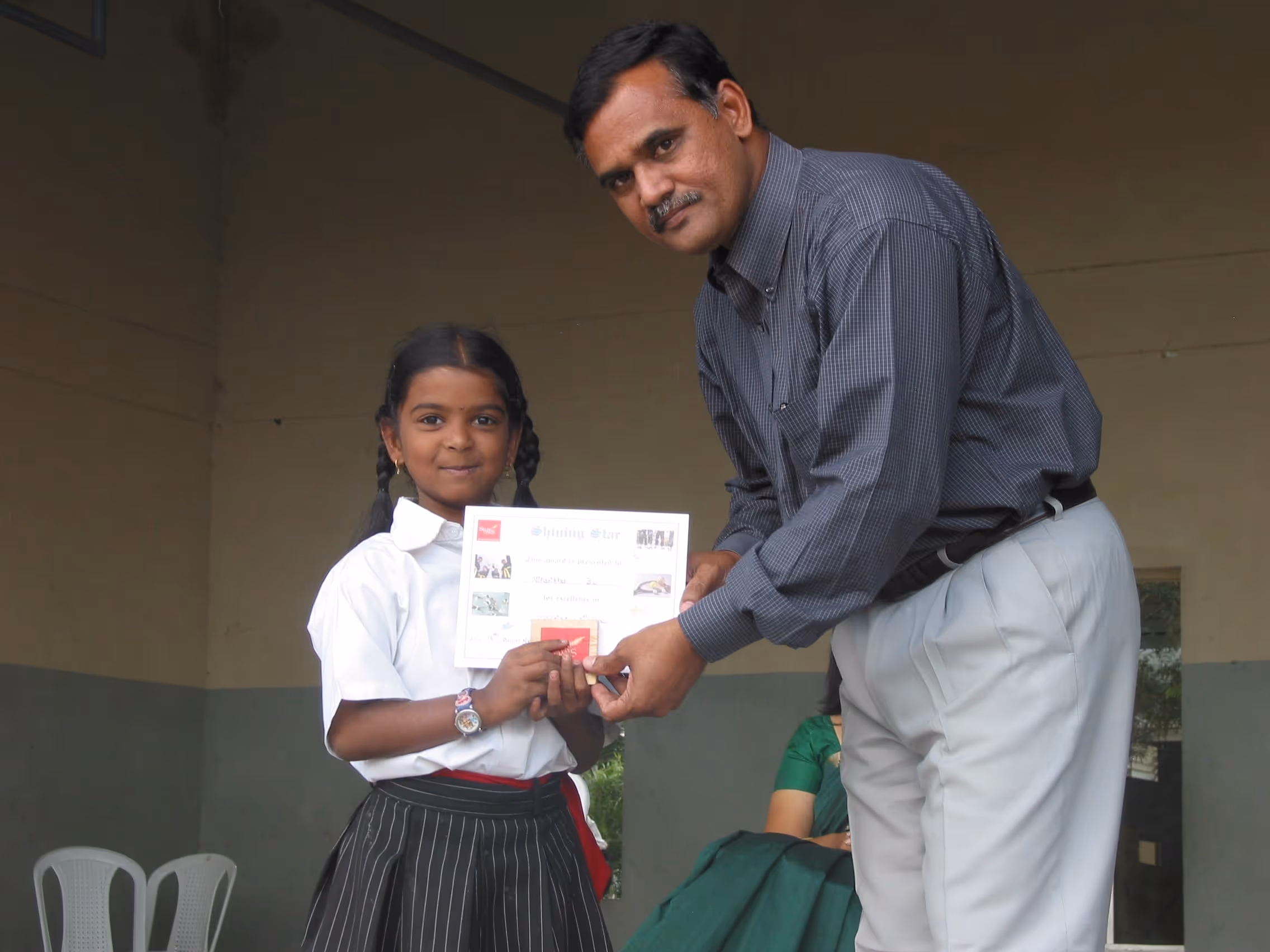 A young girl in a school uniform receives a certificate from a man in a gray shirt and light trousers indoors.