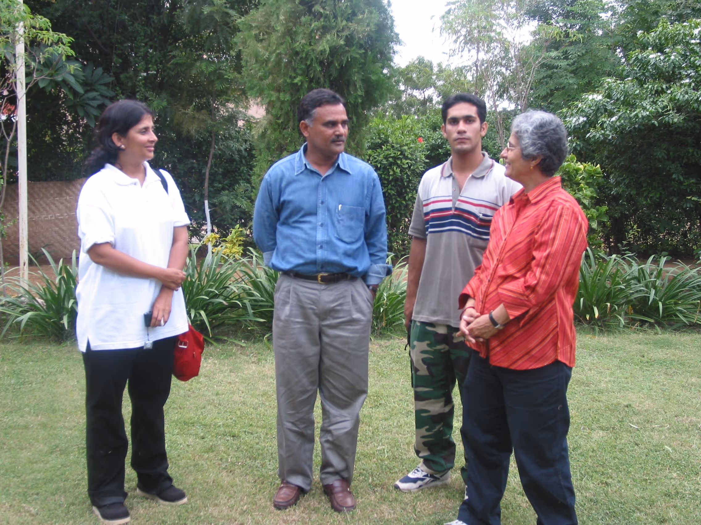 Four adults standing on grass in a garden with green plants and trees in the background.