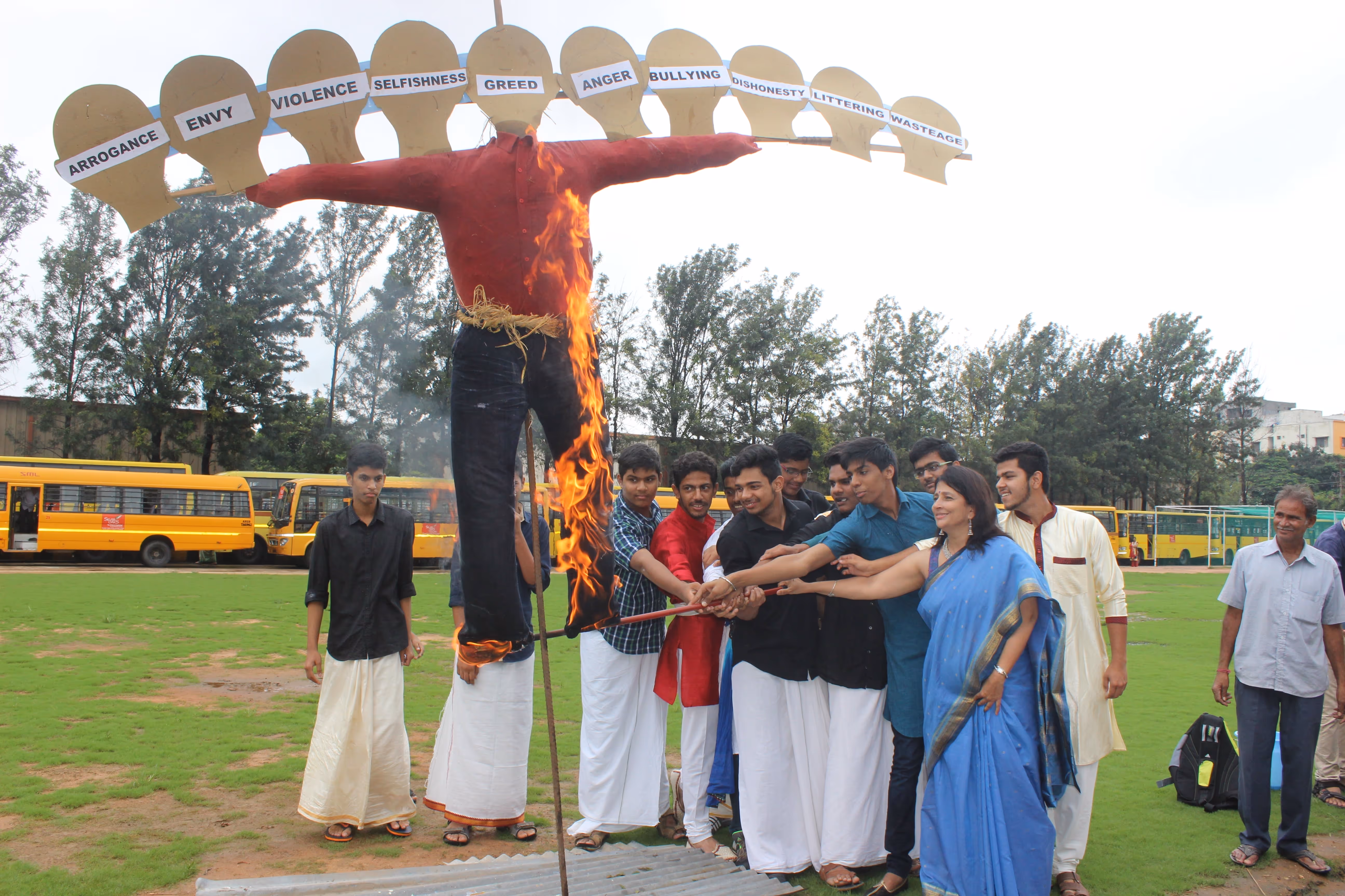 Group of people lighting a burning effigy labeled with negative traits such as arrogance, envy, violence, selfishness, and greed in an outdoor setting.