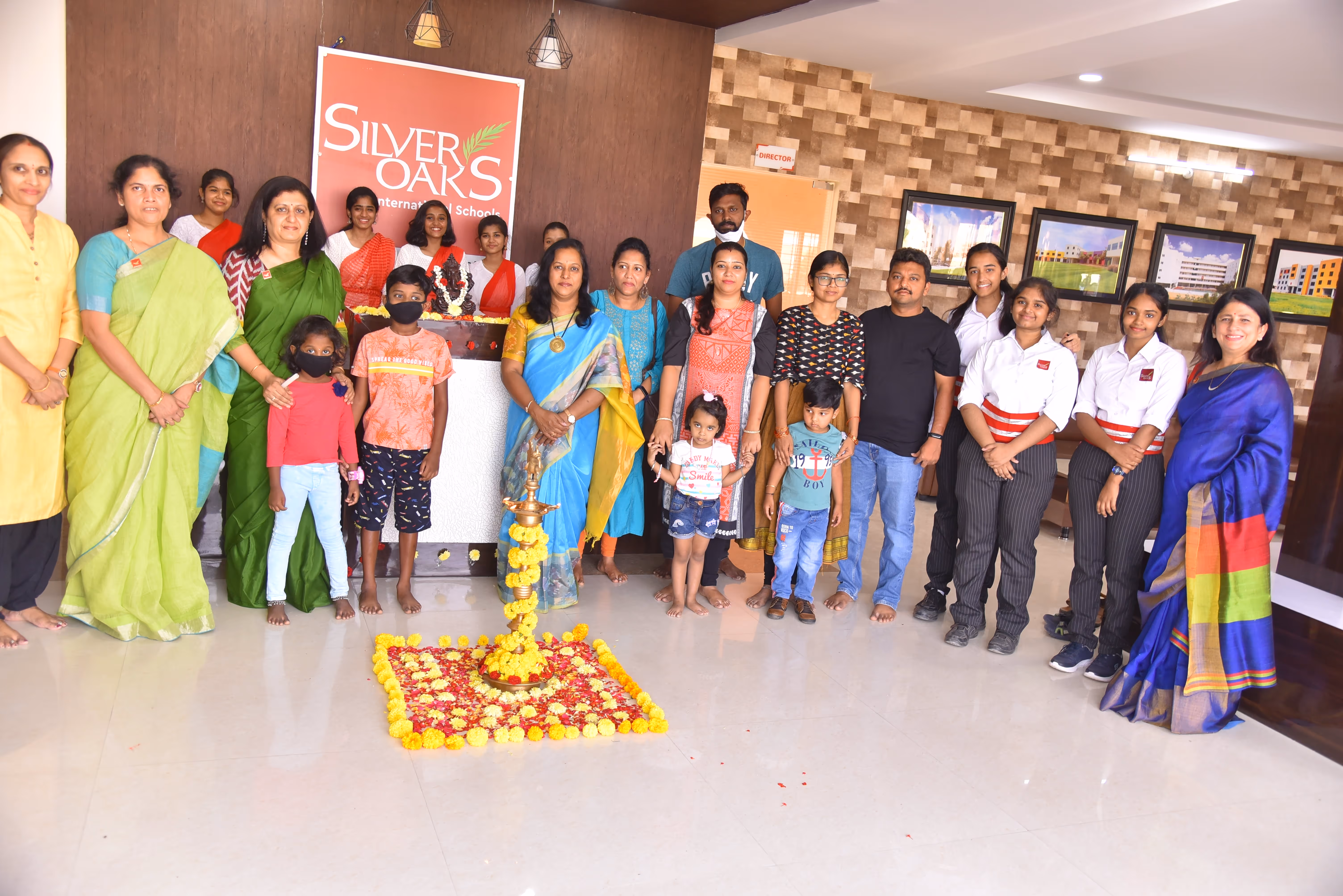Group photo of diverse adults and children standing indoors near a floral arrangement and a Silver Oaks International Schools sign.