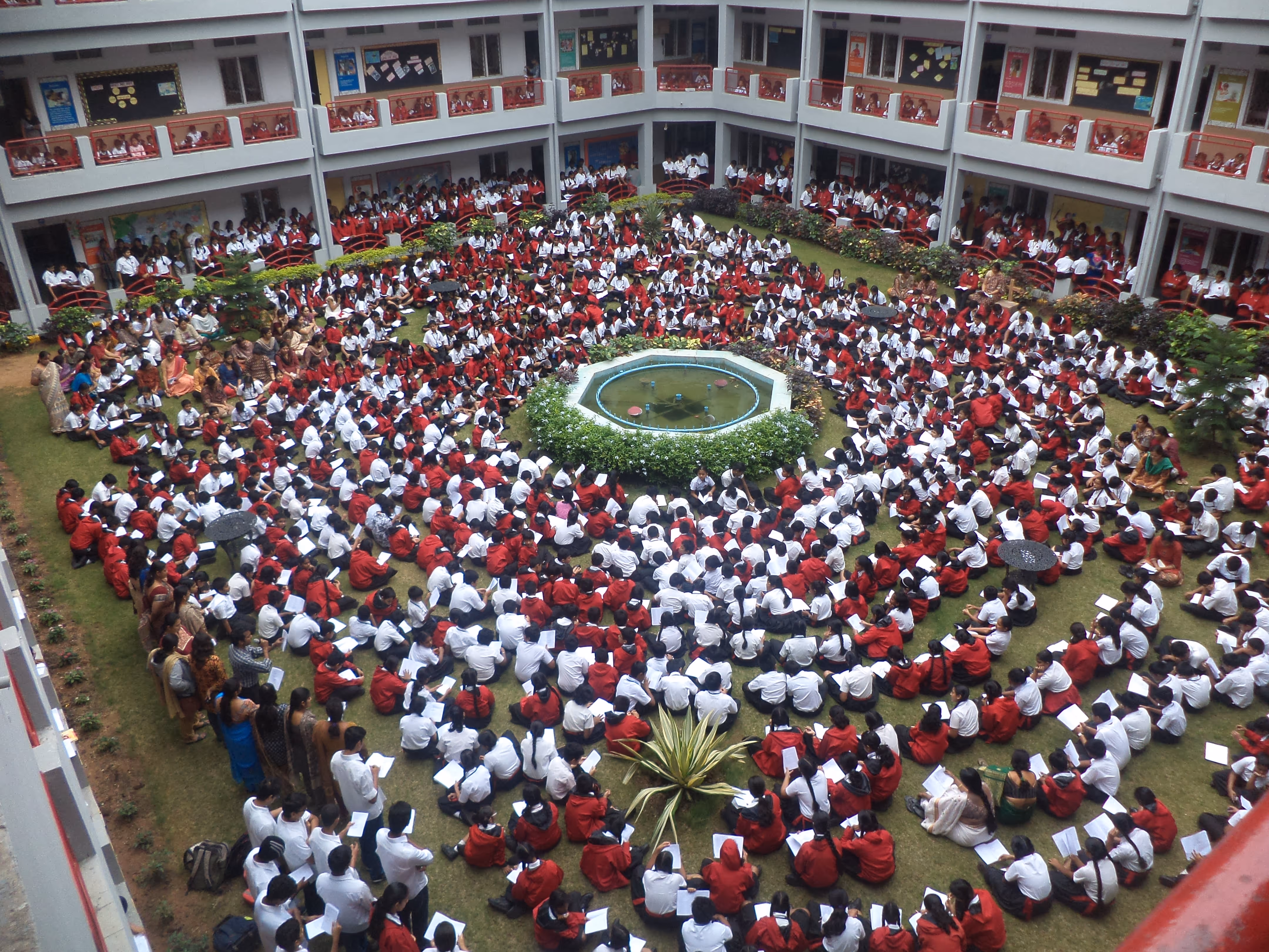 Large group of students in white shirts and red uniforms seated in concentric circles around a central fountain in a school courtyard.
