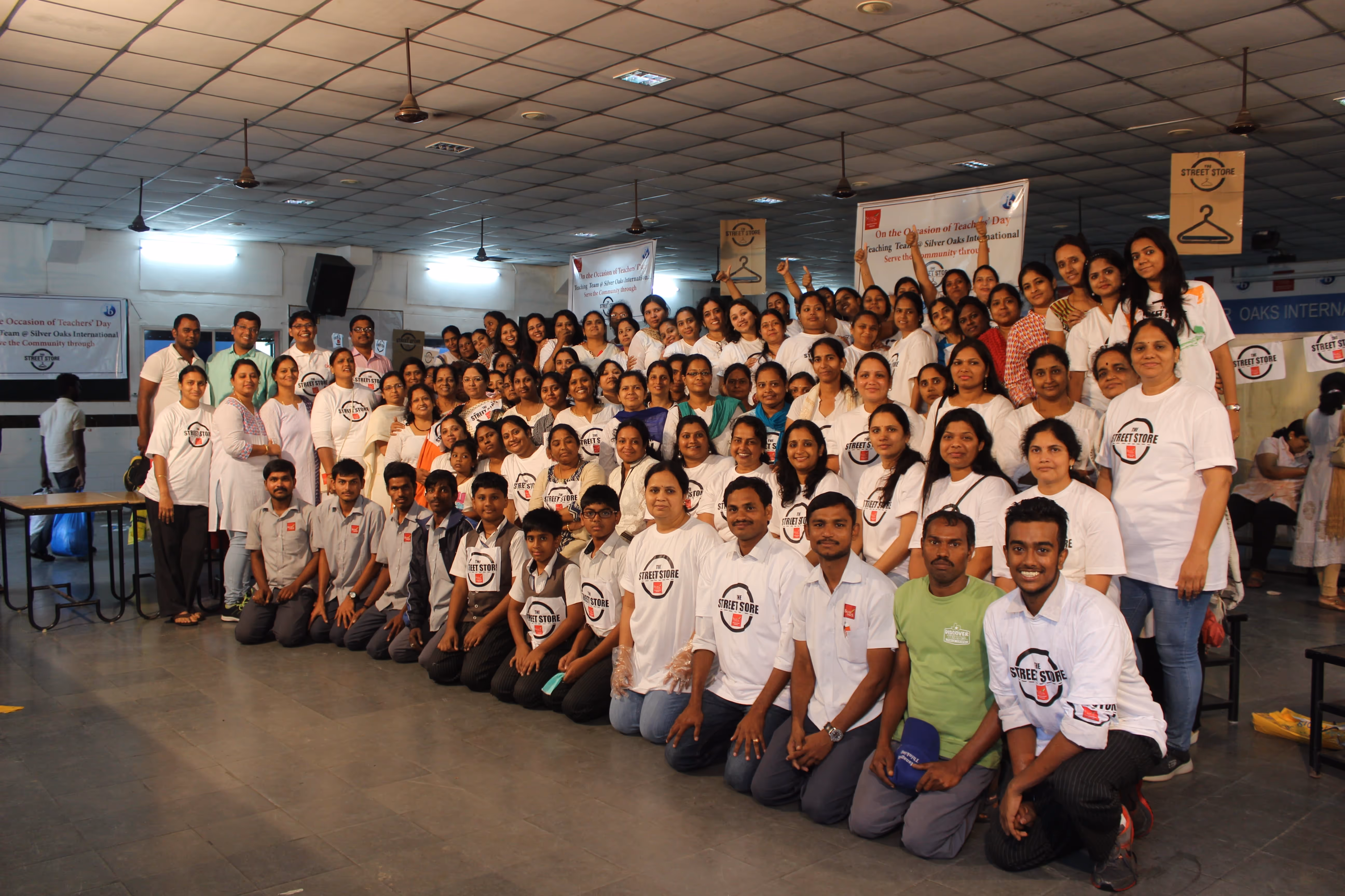 Large group of people posing indoors wearing 'The Street Store' t-shirts in front of banners celebrating Teachers' Day.
