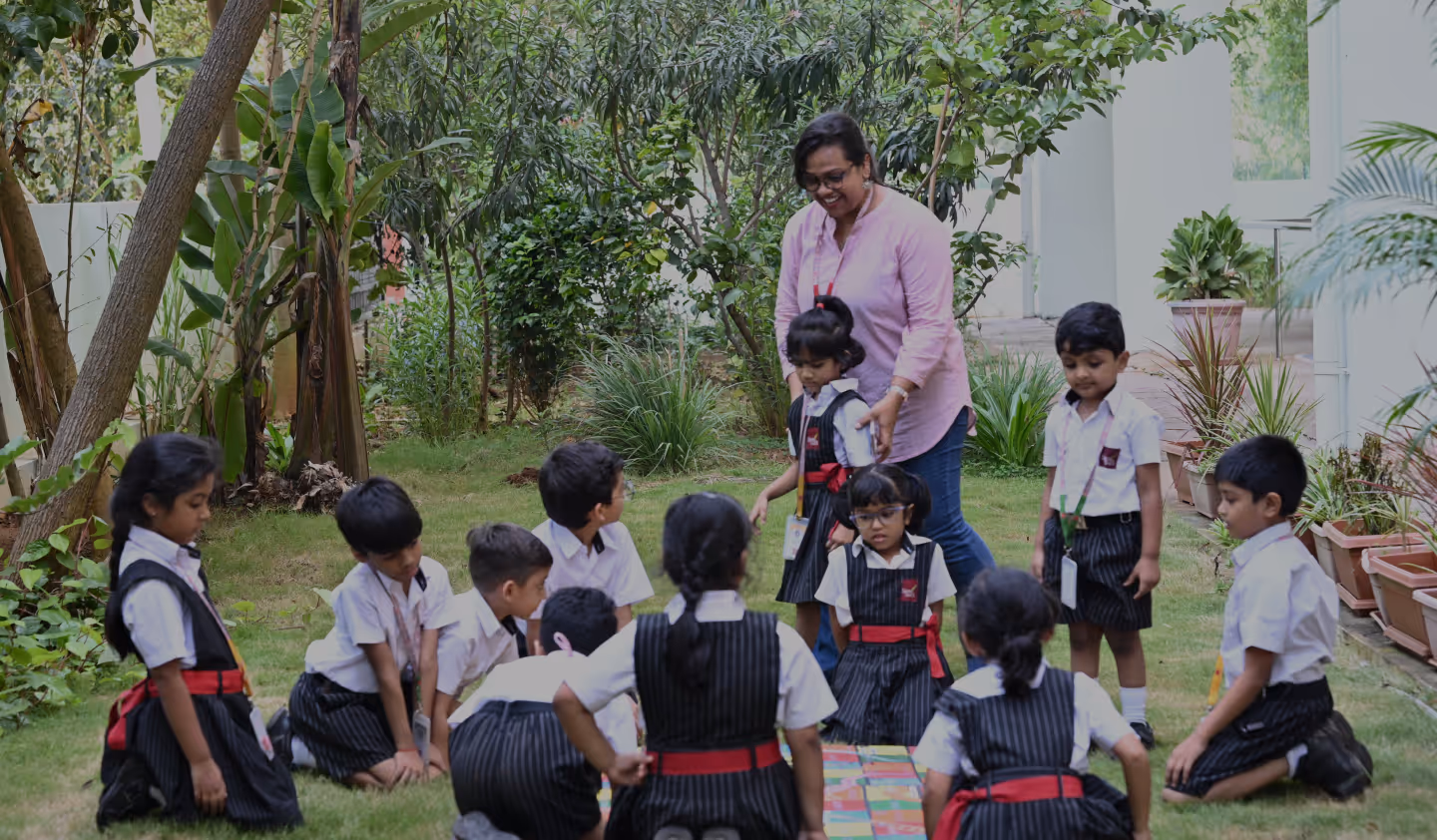 A woman with glasses and a pink top guides a group of young children in school uniforms sitting on grass near plants and trees.