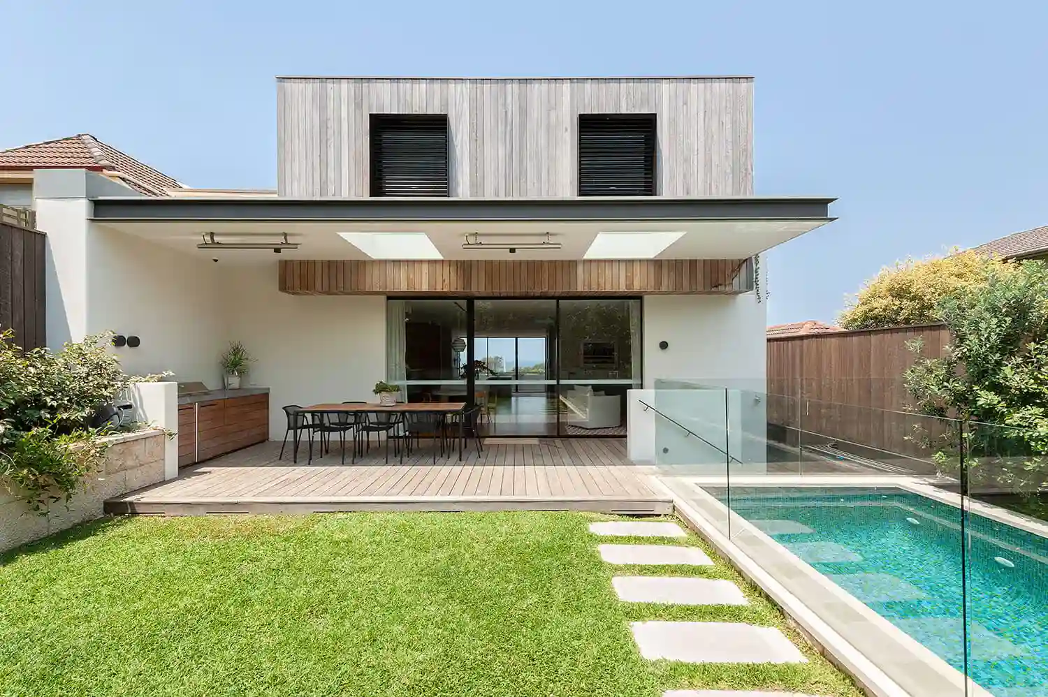 Modern backyard with wooden deck dining area, green lawn, and glass-fenced pool beside house.