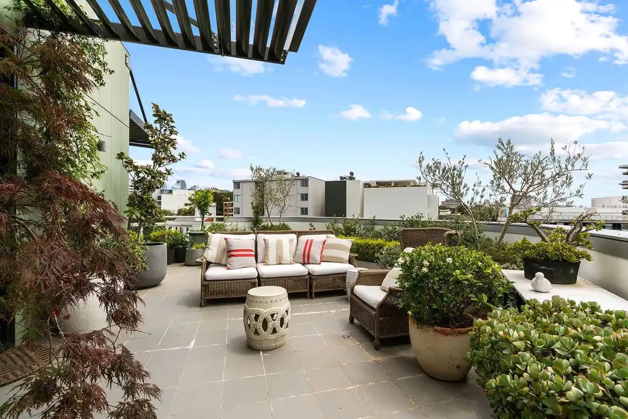 Rooftop patio with wicker sofa, armchair, cushions, plants, and city buildings under blue sky.
