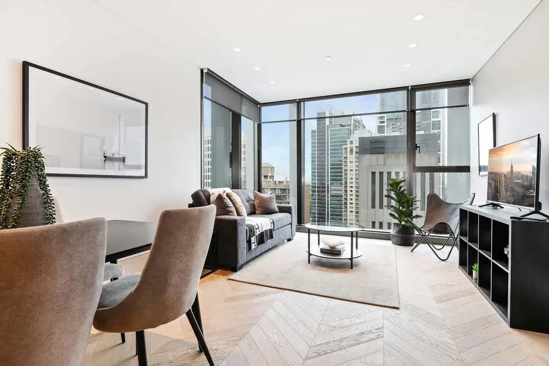 Modern living room with light wood chevron flooring, grey sofa with cushions, round glass coffee table on beige rug, black TV stand with flat screen TV, two beige dining chairs and table on the left, large floor-to-ceiling windows showing cityscape.