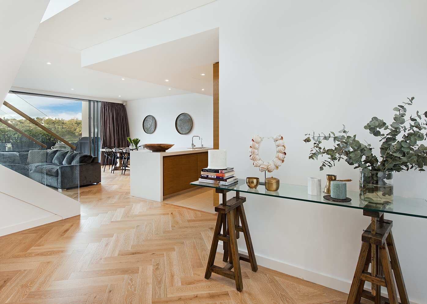 Modern living room with glass walls, a TV, two armchairs, sofa with cushions, and plants inside and outside.
