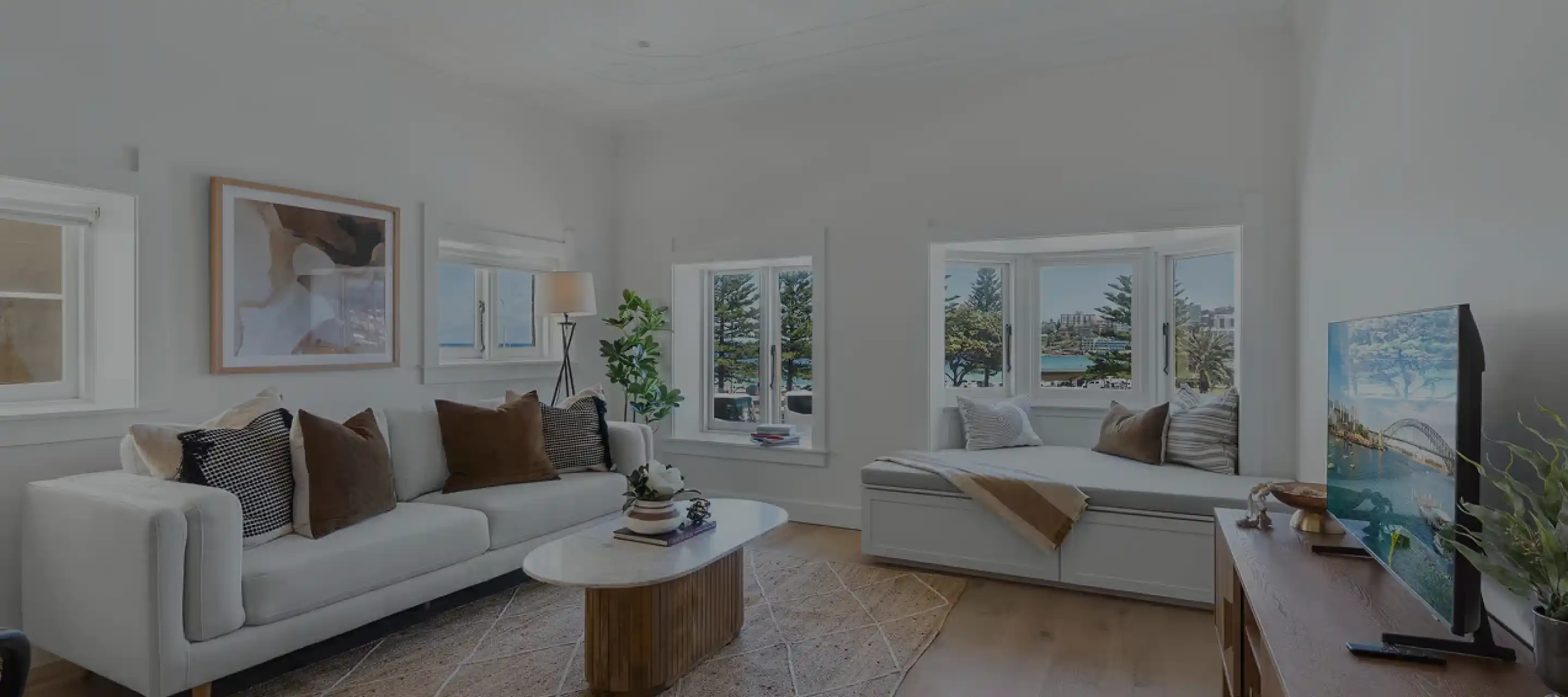Bright living room with white sofa adorned with brown and black cushions, a wooden coffee table with decorative items, and a window seat with pillows and a throw blanket. Large windows offer a view of trees and water. A TV on a wooden stand shows an image of a cityscape with a bridge over water. Light hardwood floors and white walls complete the space.