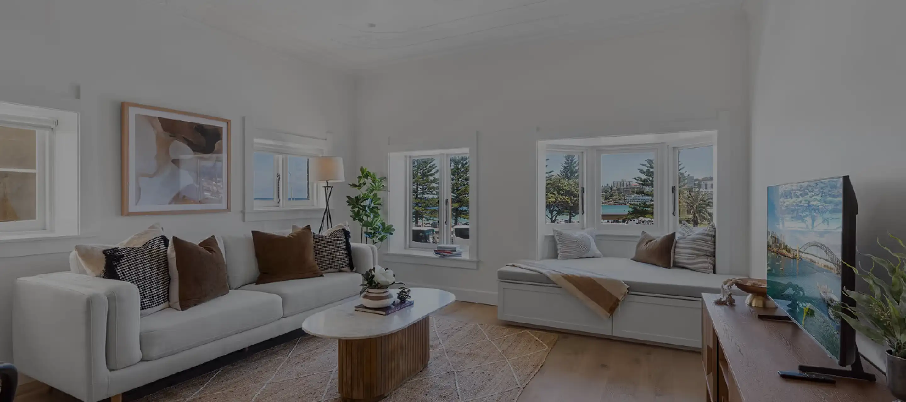 Bright living room with white sofa adorned with brown and black cushions, a wooden coffee table with decorative items, and a window seat with pillows and a throw blanket. Large windows offer a view of trees and water. A TV on a wooden stand shows an image of a cityscape with a bridge over water. Light hardwood floors and white walls complete the space.