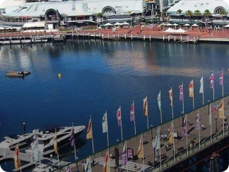 View of a waterfront marina area with yachts docked along piers, a row of colorful flags lining the walkway, and industrial buildings in the background.