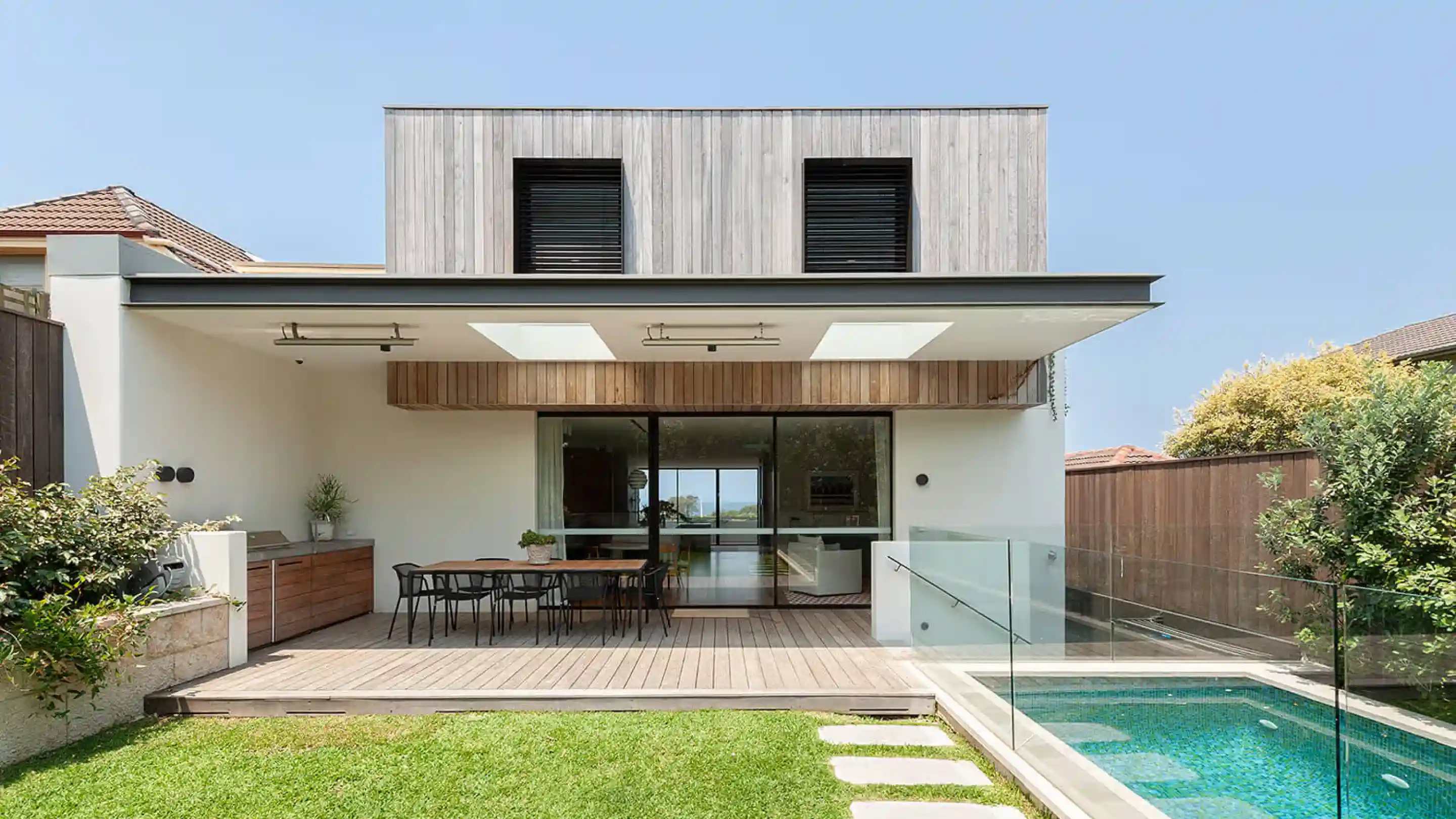 Modern two-storey house with a wooden deck patio, outdoor dining table with chairs, built-in outdoor kitchen, glass safety fence around a tiled swimming pool, green lawn with stepping stones, and wooden panel siding on the upper floor under a clear blue sky.