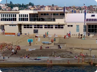 Clovelly Beach with swimmers and sunbathers on sandy shore and concrete steps, beige buildings in background, and cityscape beyond under clear sky.