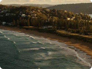 Coastal landscape with gentle ocean waves rolling onto a sandy beach bordered by dense trees and houses on a hillside under a cloudy sky at sunset.