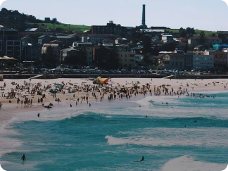Crowded beach with many people sunbathing, swimming, and surfing along the coastline with buildings and greenery in the background under a clear sky.
