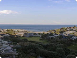 Coastal view of a beach with calm ocean waves, surrounded by greenery and residential houses under a clear blue sky with some clouds.