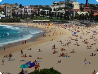 Crowded sandy beach with many people sunbathing and swimming, colorful umbrellas near the shore, and multi-story buildings and greenery in the background under a clear sky.
