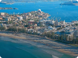 Aerial view of a coastal city with buildings near a sandy beach and calm ocean waters, with boats visible on the water.