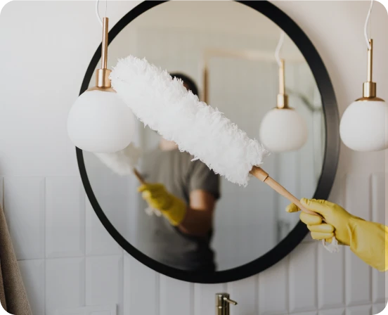 Person wearing yellow gloves using a fluffy white duster to clean a modern round mirror in a bathroom with white tiled walls and hanging pendant lights.