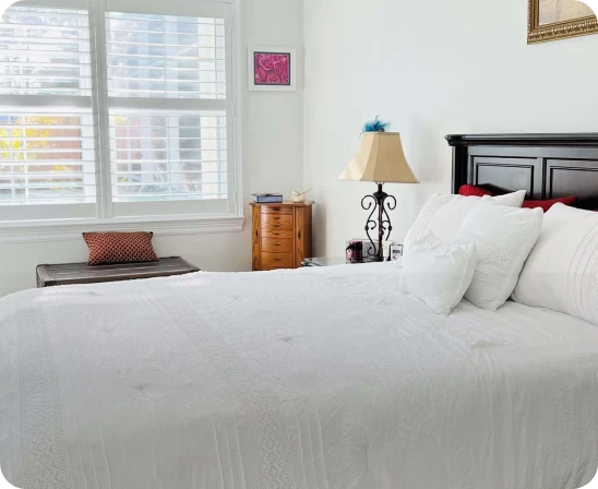 Bright bedroom with a large bed covered in white bedding and multiple pillows, a dark wooden headboard, a bedside table with a decorative lamp, a wooden chest by the window with closed shutters, and light walls with minimal decoration.