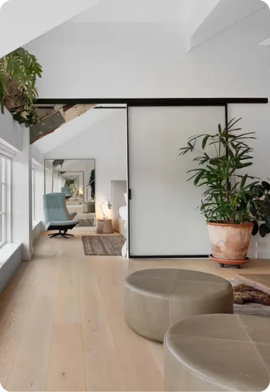Modern interior with light wooden floor, two round beige ottomans, a large potted green plant, a sliding door, and a light blue lounge chair near windows with natural light.