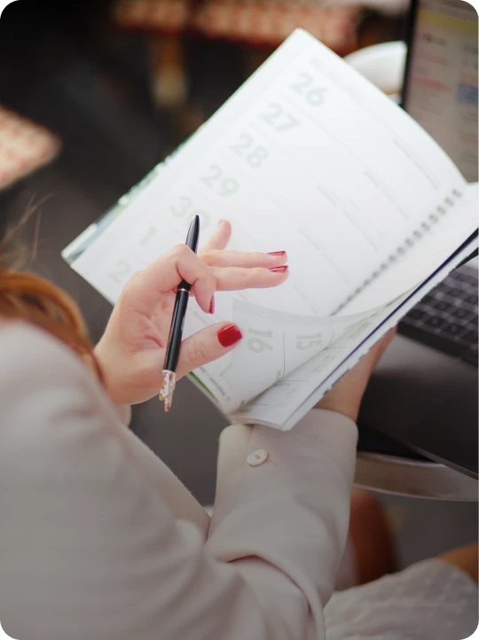 Person with red nail polish flipping pages in a ring-bound planner or calendar while holding a black pen and looking at a laptop.
