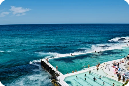 Ocean pool beside the sea with people swimming and lounging around on a sunny day with blue sky and scattered clouds.