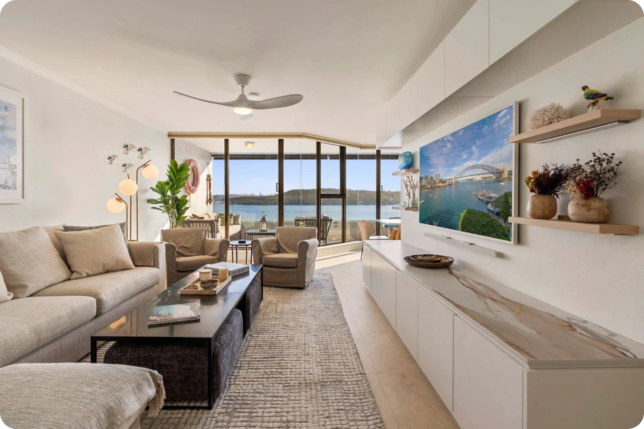 Modern bedroom with king bed, blue bench, armchair, balcony, and ocean view through floor-to-ceiling glass.