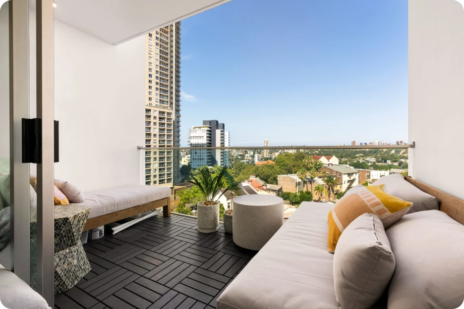 Outdoor balcony with rattan sofa, armchair, cushions, potted plants, and city buildings under blue sky.