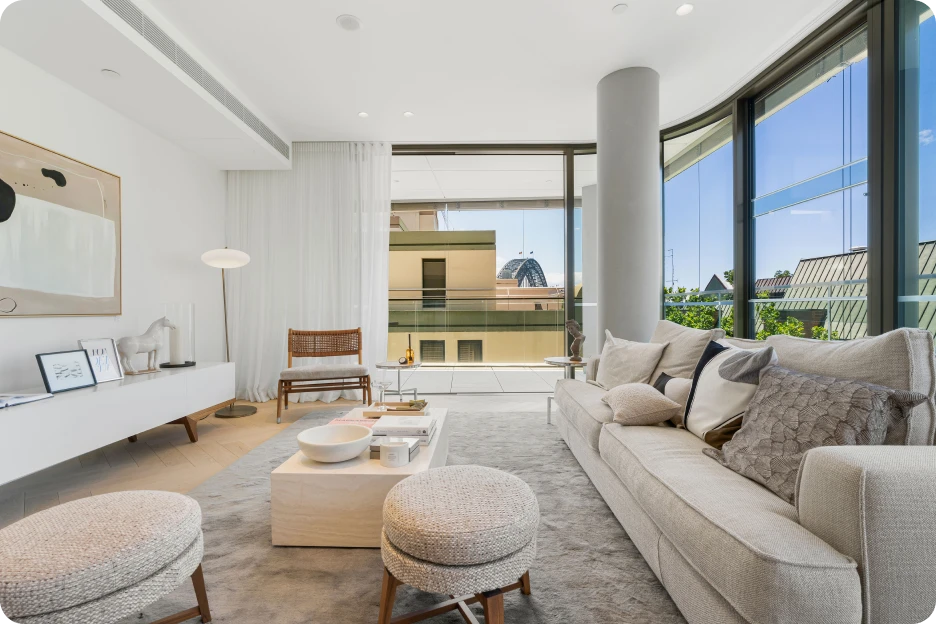 Bright living room with white sofa, window seat, wooden table, TV, and windows showing trees and water.