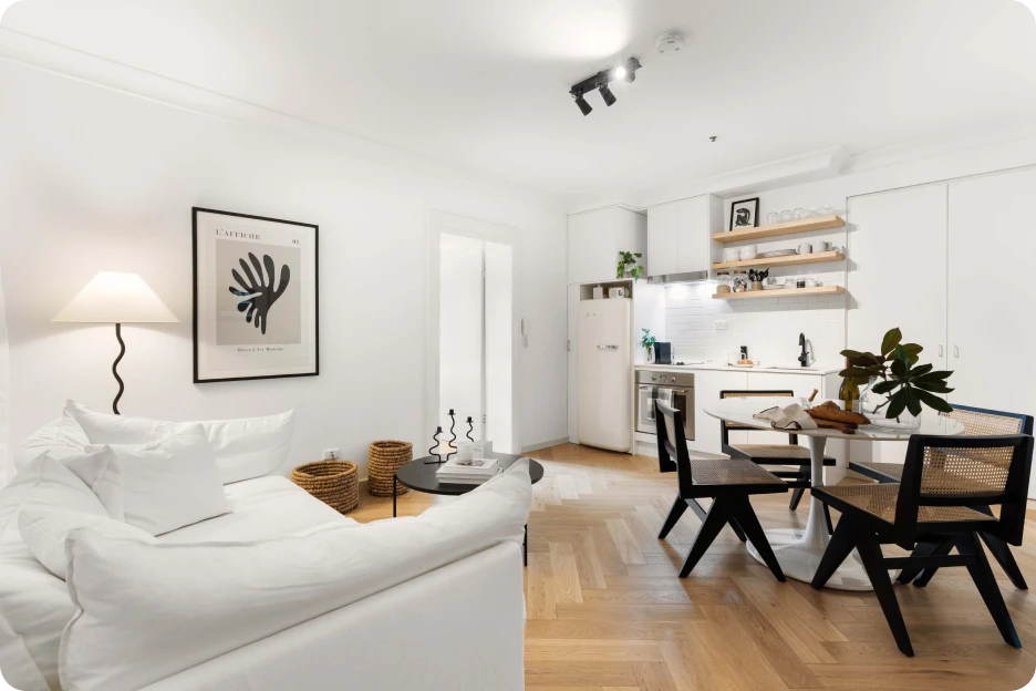 Bright modern living room with grey sofa, dining table, bookshelf, and large windows with city views.