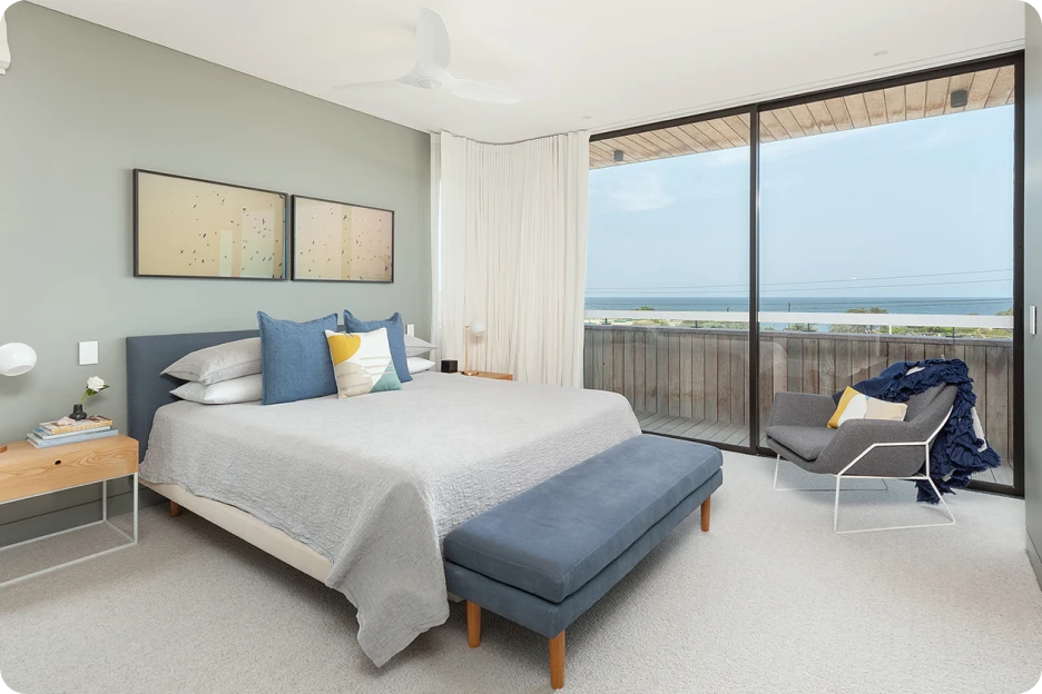 Modern bedroom with king bed, blue bench, armchair, balcony, and ocean view through floor-to-ceiling glass.