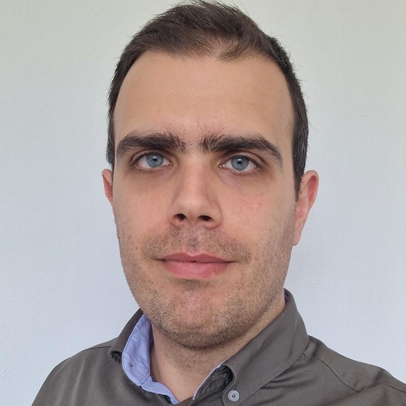 Close-up portrait of a man with short dark hair, blue eyes, and light stubble wearing a grey collared shirt with a blue inner collar against a plain light background.