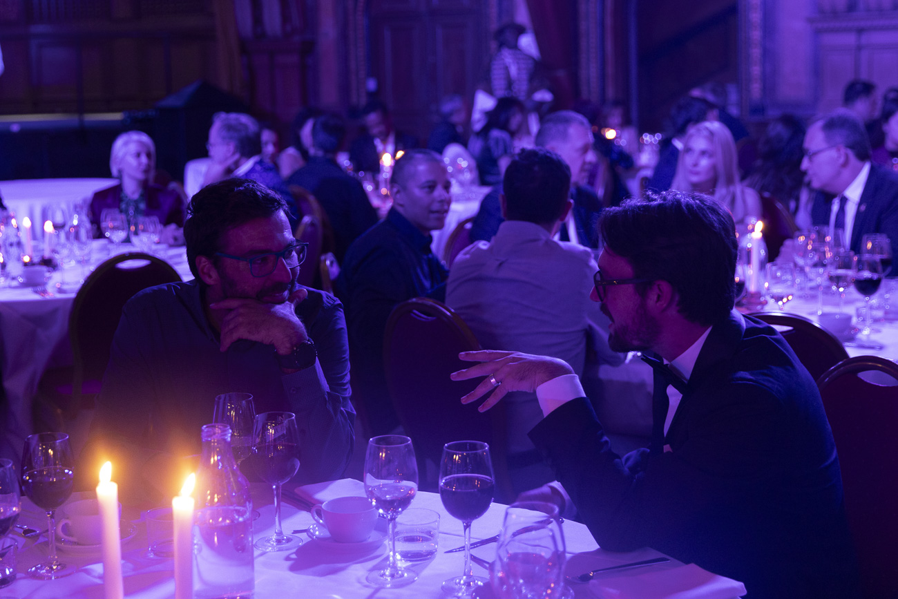 Two men in conversation at a formal dinner with candlelit tables and guests in the background.