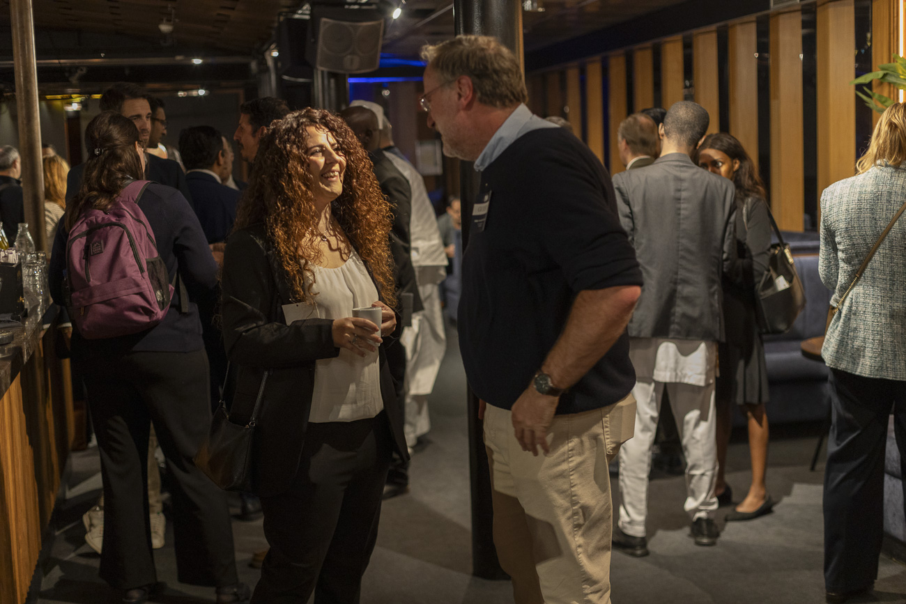 A woman with curly hair holding a cup laughs while talking to a man wearing glasses in a crowded indoor social gathering.