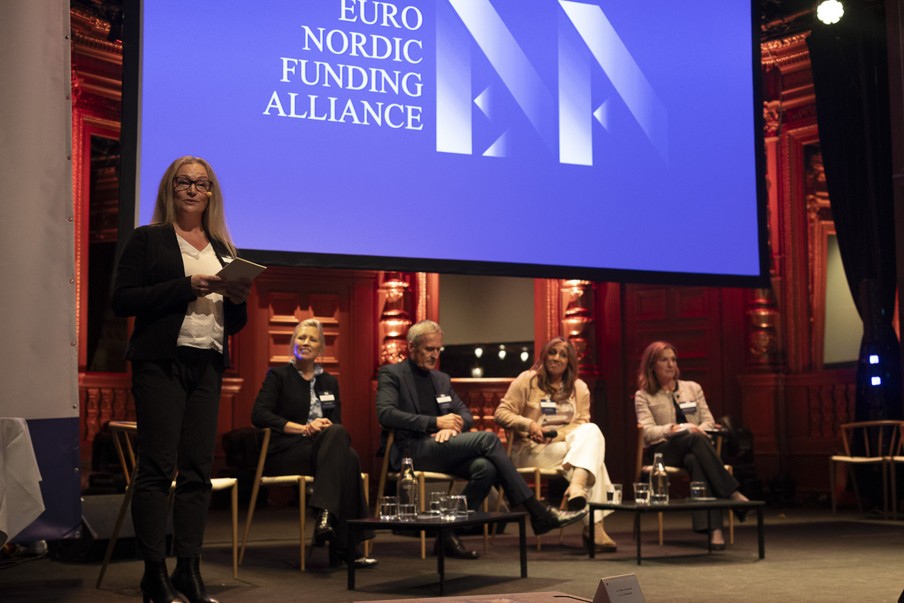 A woman speaking on stage holding notes while four panelists sit behind her under a screen displaying Euro Nordic Funding Alliance.