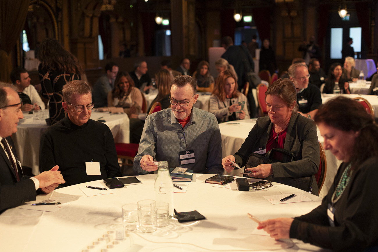 People seated around a round table at a formal indoor event, engaging in discussion and looking at papers.