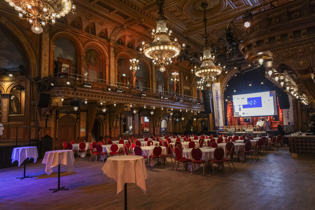 Ornate event hall with chandeliers, round tables with white tablecloths and red chairs, and a stage with a large screen for a business summit.