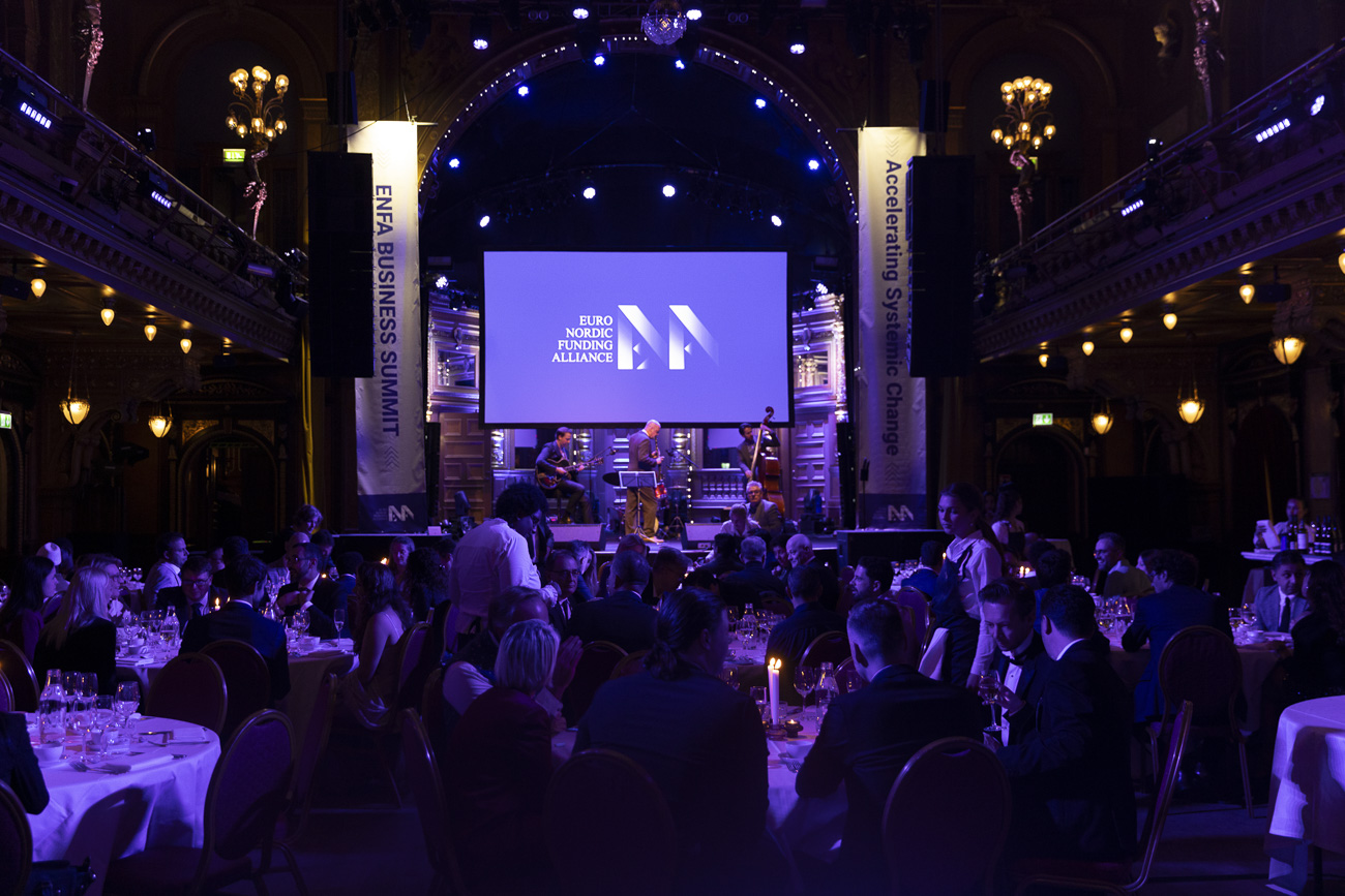 People seated at round tables with white tablecloths in a dimly lit hall watching a live jazz band perform on stage with a screen displaying 'Euro Nordic Funding Alliance'.