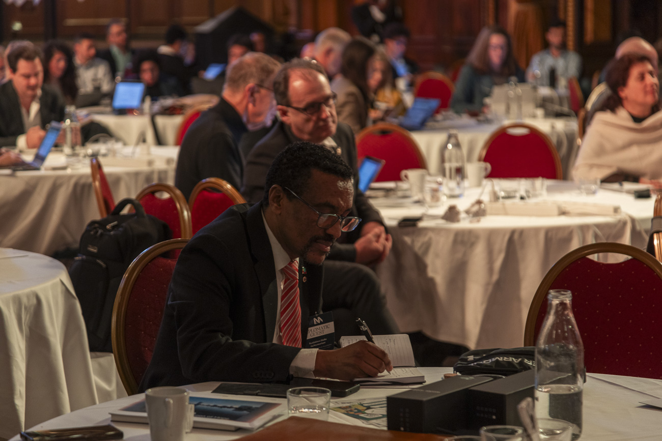 Man in a suit and red striped tie writing in a notebook at a round table during a conference.