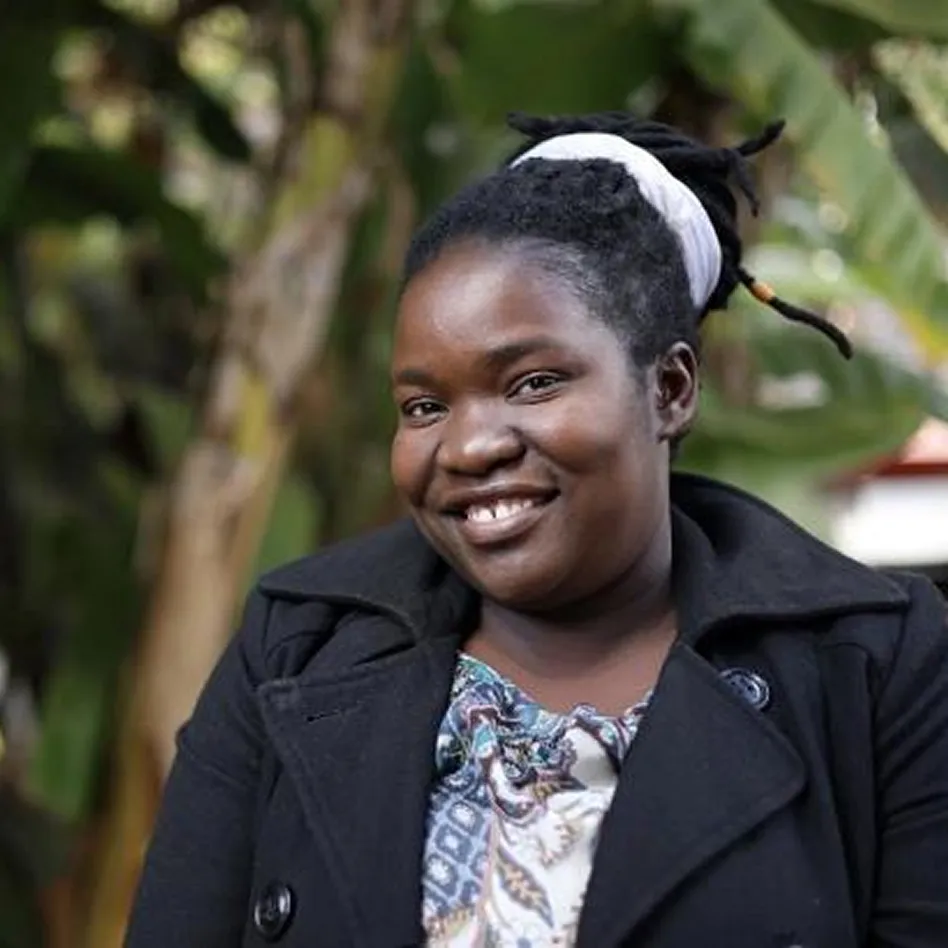 Smiling woman with dark braided hair tied with a white cloth, wearing a patterned top and black coat, outdoors with green foliage background.