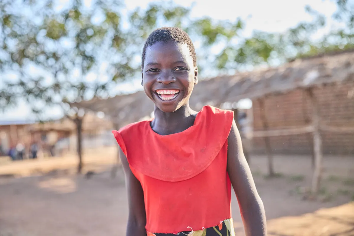 Smiling young girl wearing a red top standing outdoors with trees and a wooden structure in the background.