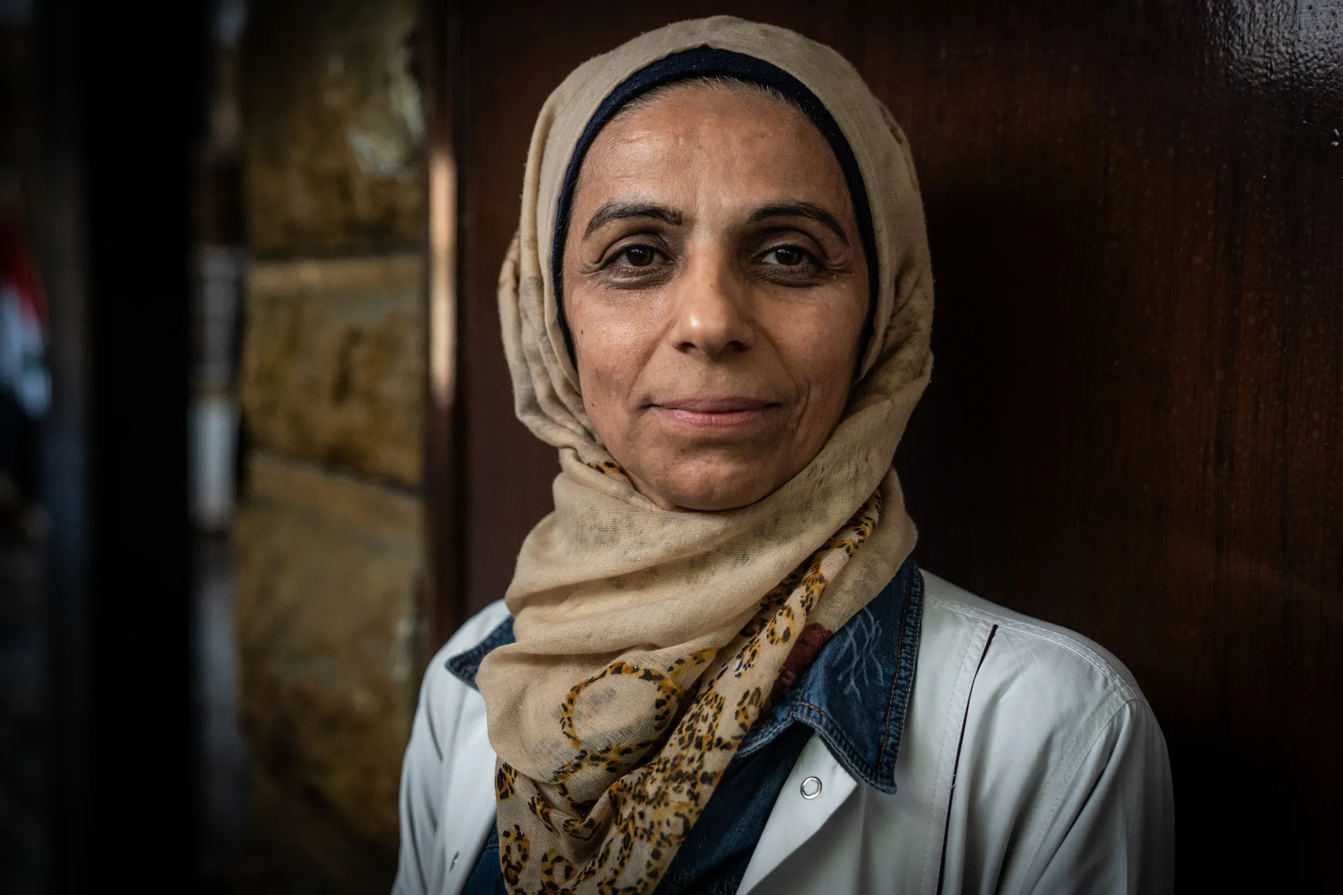Woman wearing a beige patterned hijab and white coat, standing against a wooden wall.