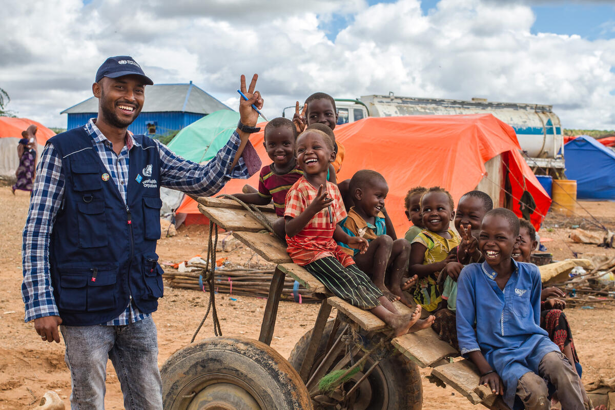 Smiling aid worker with a World Food Programme vest poses with joyful children sitting on a wooden cart in a camp with tents.